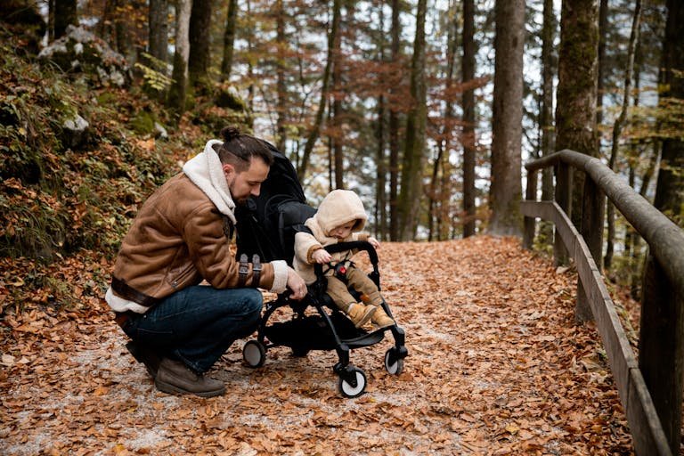 A father kneels beside his toddler in a stroller, amidst a scenic autumn forest path.