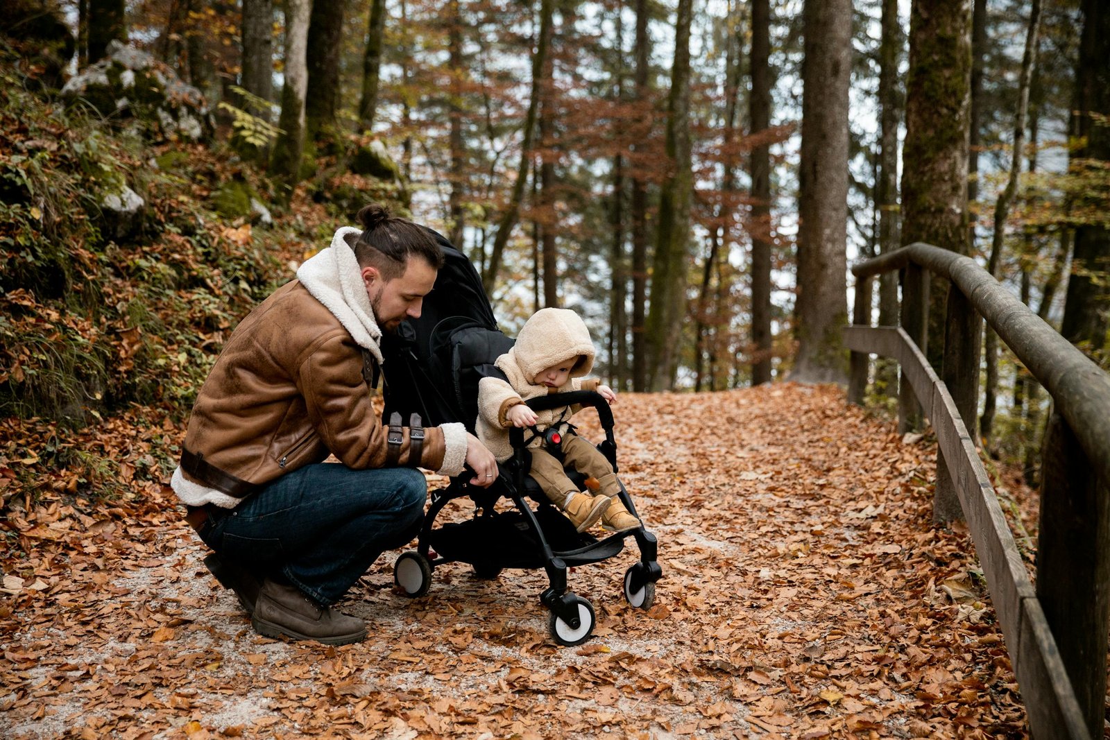 A father kneels beside his toddler in a stroller, amidst a scenic autumn forest path.